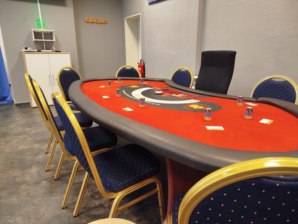Leerer Pokerraum mit rotem Tisch, blauen Stühlen und Kartenstapeln.