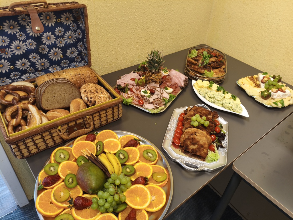 Verschiedene Speisen auf Tischen, mit Brotkorb, Fleisch, Salat, Obst und einem blauen Blumenkorb.