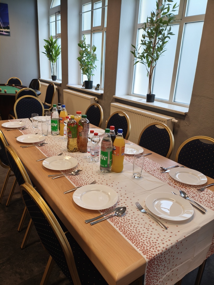 Ein gedeckter Tisch mit Getränken, Tellern und Besteck in einem Raum mit Pflanzen in den Fenstern.