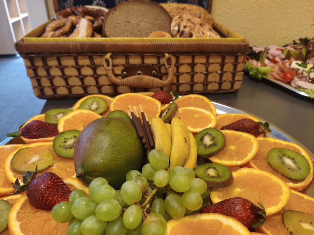 Ein Korb mit Gebäck, im Vordergrund Obstteller mit Trauben, Orangen, Erdbeeren und mehr.